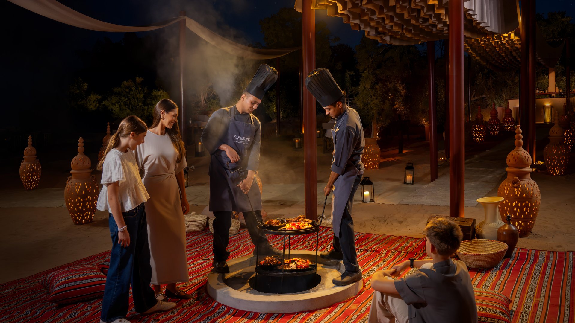 Two chefs pulling Zarb bbq meals from a ground stove surrounding by a family of four in a bedouin seating setup