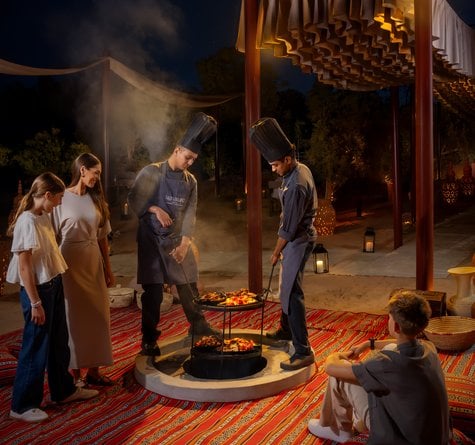 Two chefs pulling Zarb bbq meals from a ground stove surrounding by a family of four in a bedouin seating setup